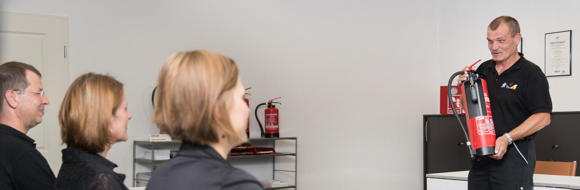 Ein Ausbilder hält einen Feuerlöscher in der Hand und erklärt einer Gruppe von Teilnehmern die richtige Handhabung. Die Schulung findet in einem Schulungsraum statt, im Hintergrund sind weitere Feuerlöscher und ein Zertifikat an der Wand zu sehen.